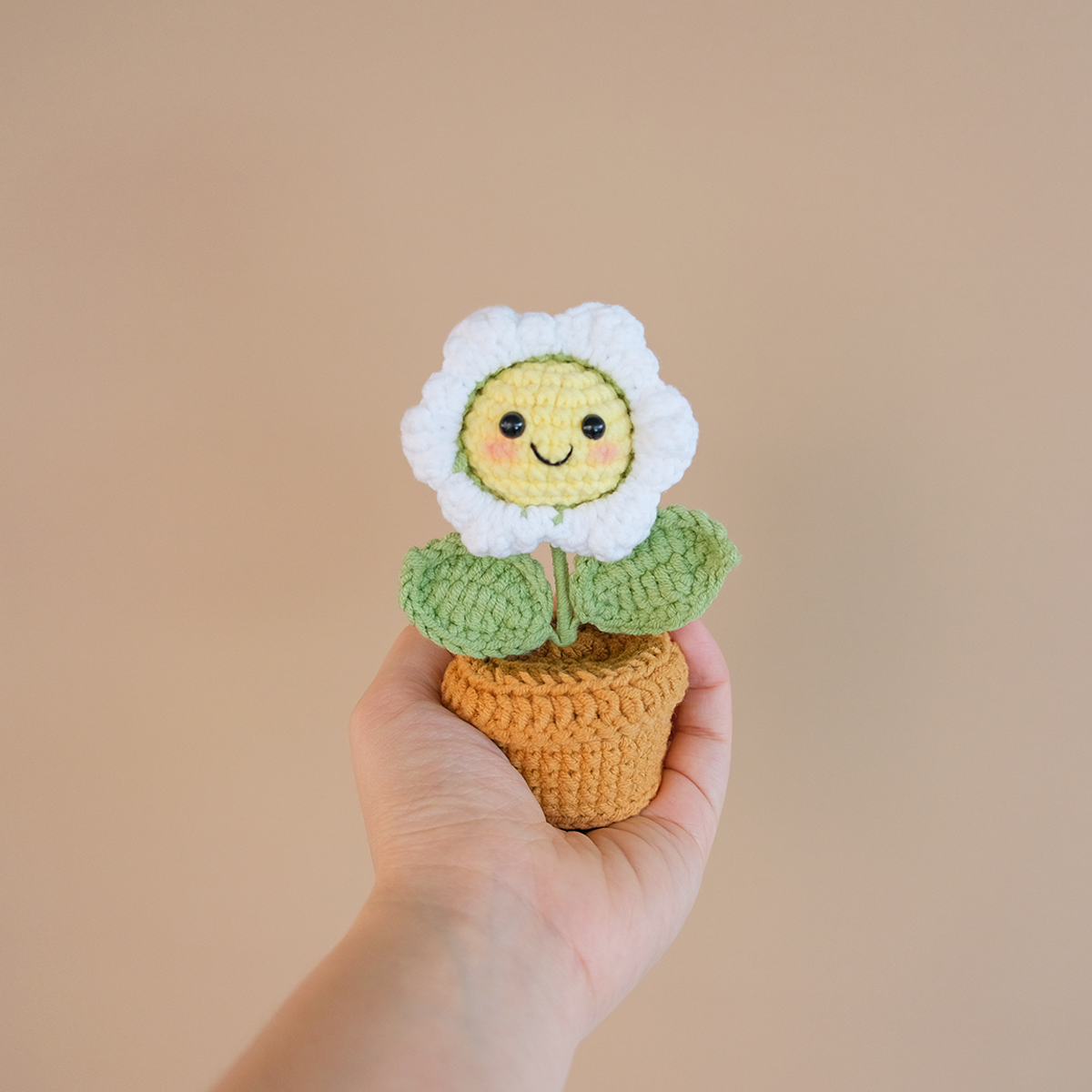 A hand holding a crocheted mini daisy flower with a smiley face in a crocheted pot.