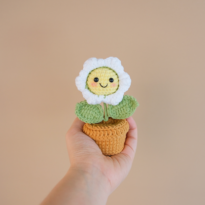 A hand holding a crocheted mini daisy flower with a smiley face in a crocheted pot.