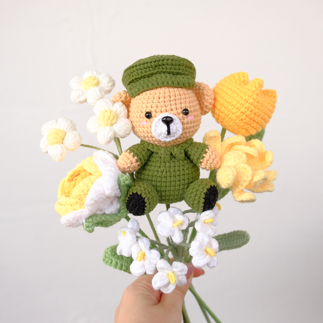 A crochet bouquet featuring a bear character with green and beige accents, surrounded by various crochet flowers