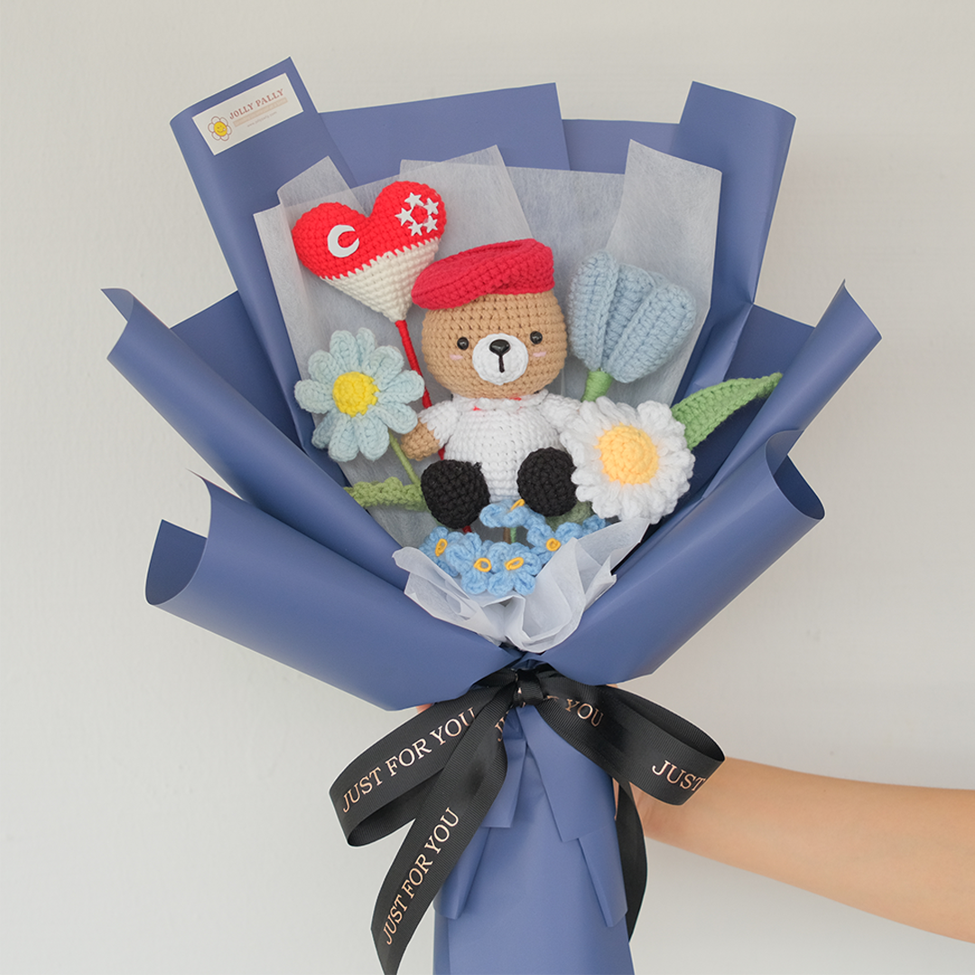 A crochet bouquet featuring a bear character with white and red beret, surrounded by various crochet flowers wrapped in blue and black ribbon