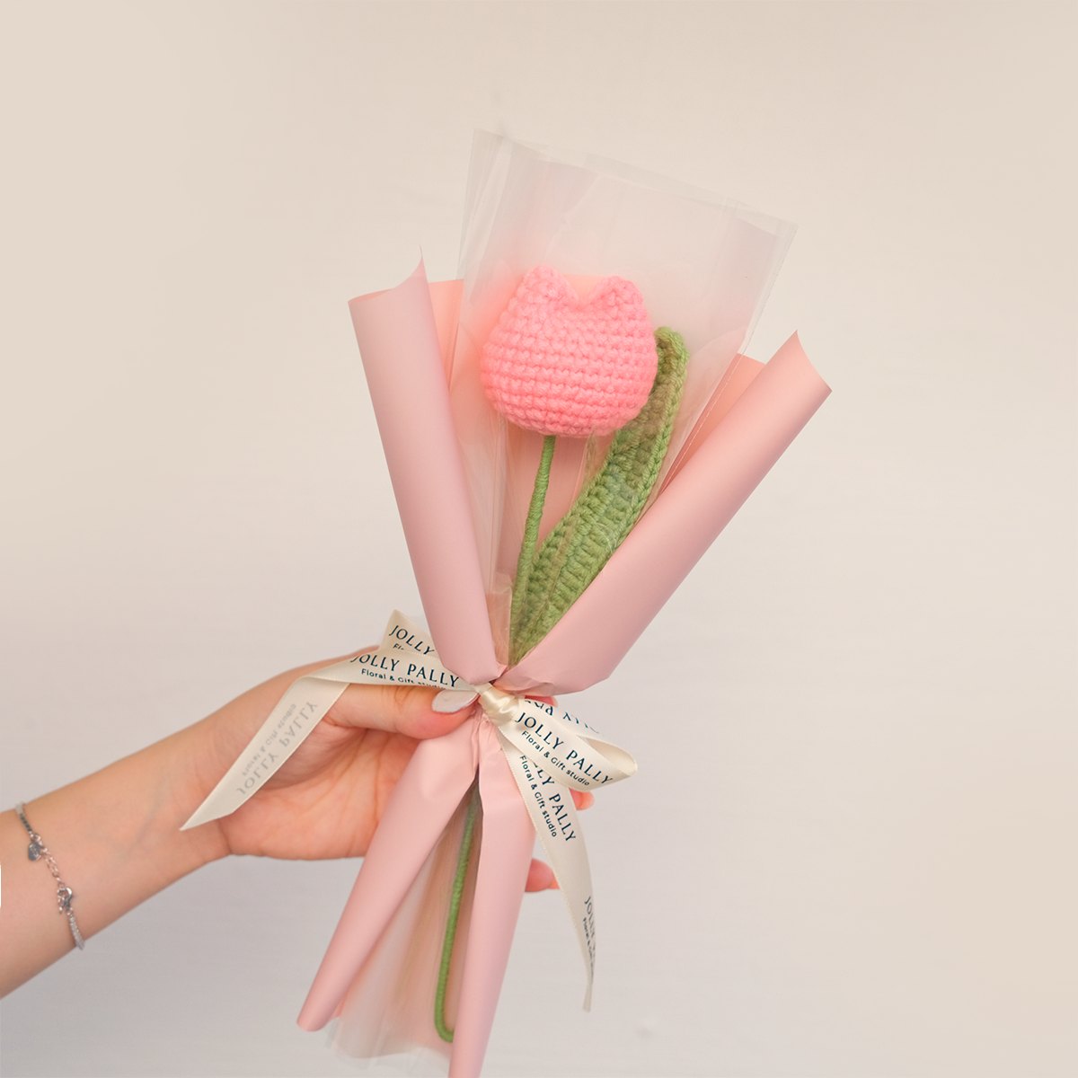 A single crochet chubby tulip flower bouquet, wrapped in pink paper with a label that reads 'Jolly Pally'. There is also a tag with a product code visible.