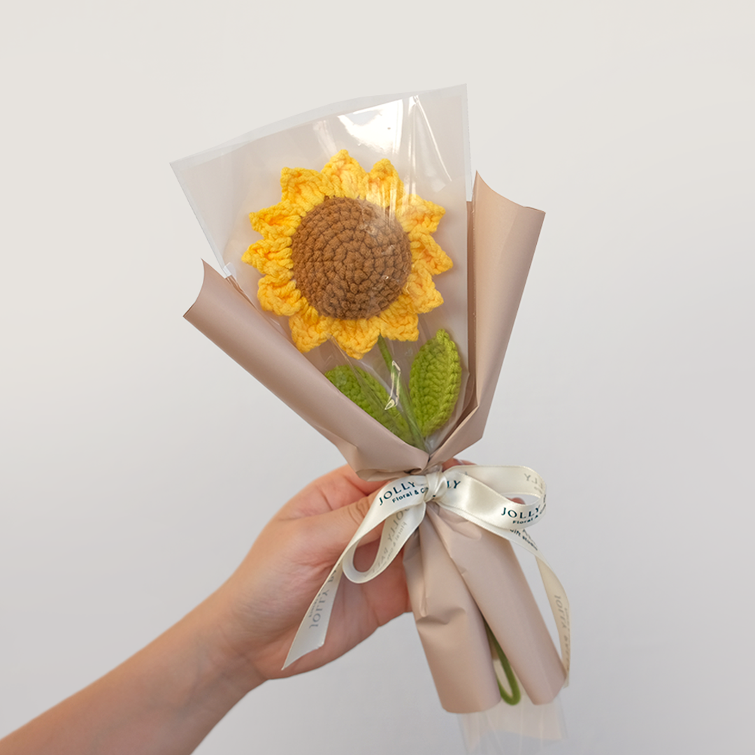 A hand holding a single crochet sunflower bouquet with brown, green, and yellow colors, wrapped in beige floral wrapping with a ribbon.